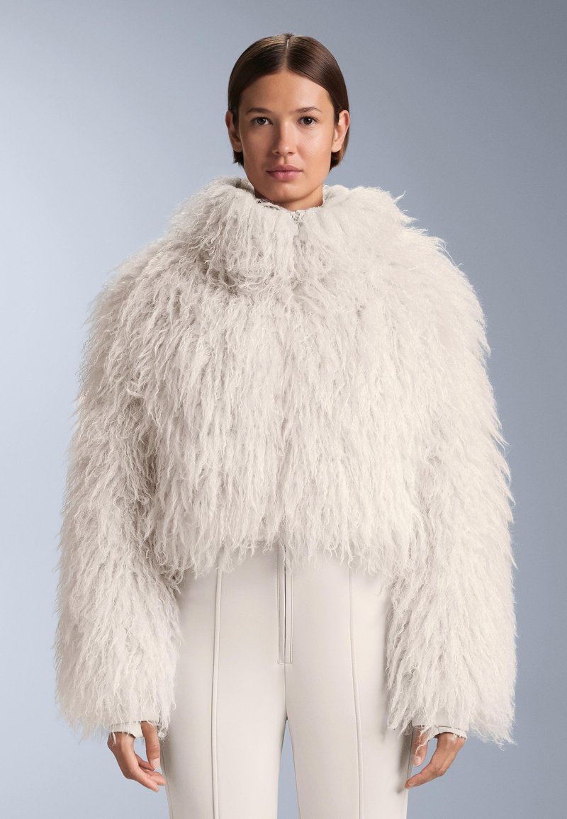 Biały, futrzany krótki płaszcz z wysokim kołnierzem i długimi rękawami. Teksturowany materiał nadaje puszysty wygląd, zestawiony z dopasowanymi jasnymi spodniami.