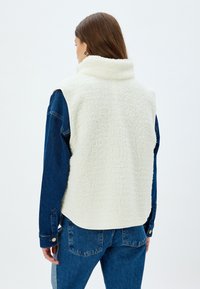 Weiße Fleece-Weste mit hohem Kragen, ärmellosem Design und strukturierter Oberfläche, über einem blauen Jeanshemd getragen, mit sichtbaren Nähten.