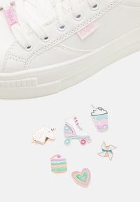 Baskets blanches à semelle plate, ornées d'un détail de logo rose. Six pin's émaillés décoratifs : licorne, patin à roulettes, boisson, gâteau, cœur et moulin à vent.