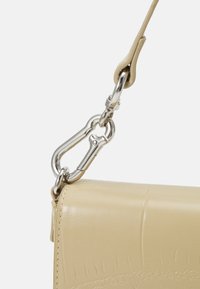 Beige nahkakotelo, jossa on sileä pinta, hopeinen metallisalonki ja hihnan kiinnitys. Huomionarvoinen ompelutyyli reunassa.