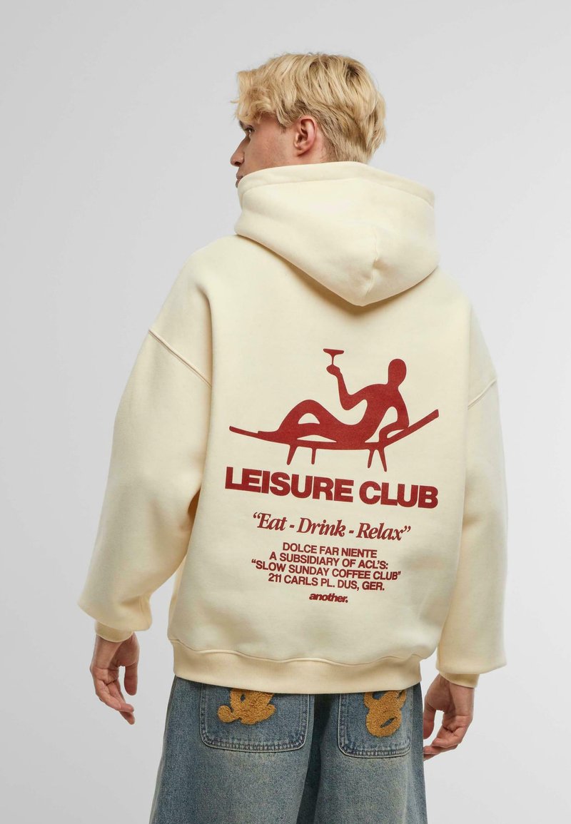 Felpa crema con stampa grafica rossa sul retro, con testo e una figura in relax. Il materiale appare morbido con una vestibilità comoda.