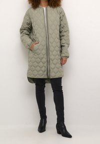 Vadderad, olivgrön långjacka med dragkedja, två sidofickor och ribbade ärmslut, ihop med mörka jeans och svarta klackstövlar.