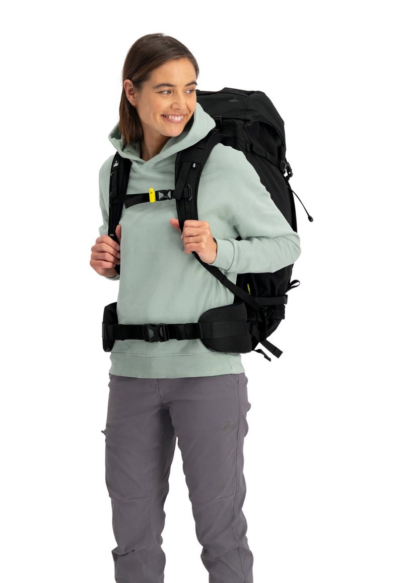 Zwarte rugzak met verstelbare banden en gele hardware. Draagbare over een lichtgroene hoodie en grijze broek. Beschikt over een gevoerde heupband.