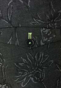 Schwarzer Stoff mit dezentem Blumenmuster, ausgestattet mit einer grünen Akzentnaht und einem schwarzen Knopf mit grünem Detail. Glatte Textur.