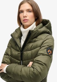 Chaqueta acolchada verde oliva con cierre de cremallera, capucha y puños acanalados. Presenta un parche de logo redondo en el brazo izquierdo y textura acolchada.