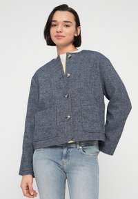 Μπλε ναυτικό, υφασμάτινο σακάκι με στρογγυλή λαιμόκοψη, κλείσιμο με κουμπιά μπροστά, δύο τσέπες με μπαλώματα και μακριά μανίκια, συνδυασμένο με ανοιχτόχρωμα μπλε τζιν.