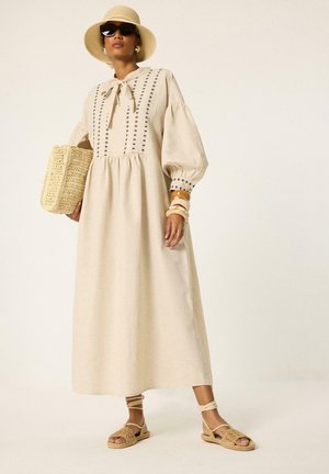 Vrouw die een beige midi-jurk met lange mouwen en zwarte borduurwerk draagt, een hoed met brede rand, zonnebril, geweven sandalen, een gevlochten tas draagt en armbanden draagt.