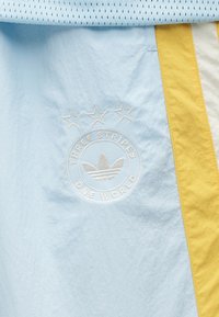 Ανοιχτό μπλε ύφασμα με κεντημένο κυκλικό λογότυπο που περιλαμβάνει τρία αστέρια και τη φράση "THREE STRIPES ONE WORLD", συμπληρωμένο με κίτρινες πλαϊνές πινελιές.