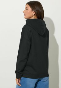 Felpa nera con cappuccio, dalla vestibilità rilassata, con polsini e cintura a costine; texture morbida, indossata con jeans blu, mostrata da dietro.
