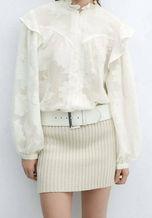 Femme portant un chemisier blanc transparent à fleurs avec des manches bouffantes, une ceinture blanche et une mini-jupe en maille côtelée beige.