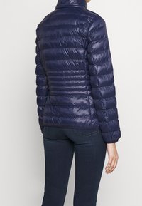 Chaqueta de puffer azul marino con un alto cuello, diseño acolchado, acabado brillante y puños elásticos, combinada con jeans oscuros.