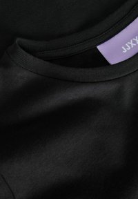 Svarte bomull t-skjorter lagd i lag, med rund hals og synlig etikett med lilla bakgrunn. Jevn tekstur, minimalistisk design.
