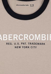 Beige t-skjorte med navy crew-hals. Fronten har fet hvit "ABERCROMBIE" tekst over mindre svart tekst, inkludert "REG. U.S. PAT. TRADEMARK."