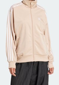 Beige džinová bunda na zip s vysokým límcem, bílý design se třemi pruhy podél rukávů a malým logem na hrudi. Hladká textura, volný střih.