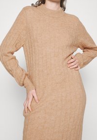 Robe en tricot côtelé beige avec un col montant et des manches longues, dotée de poches latérales et d'une coupe décontractée, fabriquée dans un matériau doux et texturé.