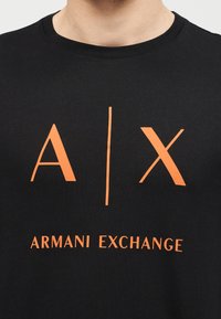 Μαύρο μπλουζάκι με πορτοκαλί λογότυπο "A | X" και κείμενο "Armani Exchange" στο κέντρο του στήθους.