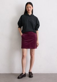 Un sweat à capuche noir avec le logo "Marc O'Polo" en relief, associé à une mini-jupe en velours côtelé bourgogne et des mocassins marron foncé avec une boucle décorative.