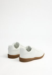 Zapatillas blancas con material sintético suave, puntera redondeada, diseño con cordones y suela de goma marrón. Tacón plano con detalles de costura mínimos.