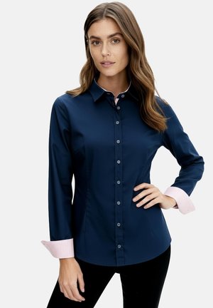 Woman in navy blue button-up shirt with pink striped cuffs poses with hand on hip against white background.
