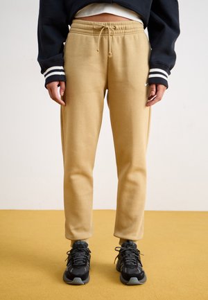 Beige bomull joggebukser med snor i midjen, tilpasset passform og ribbede mansjetter. Brukt med svarte joggesko og en mørk genser.