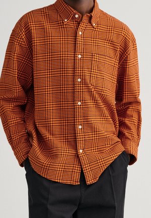 Homme portant une chemise boutonnée à motif pied-de-poule orange et noir avec poche poitrine et pantalon noir, mains dans les poches.