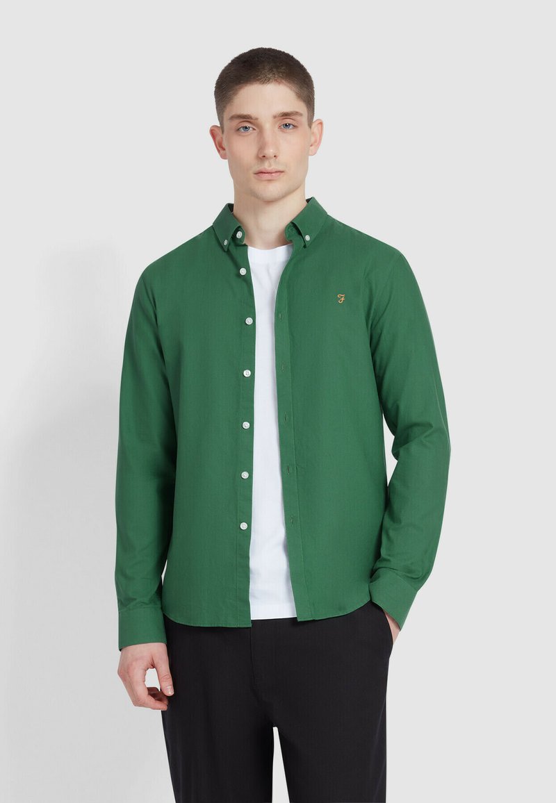 Groene button-up shirt gemaakt van gladde stof met een kraag, witte knopen en een klein geborduurd logo op de borst. Draag het over een wit shirt.