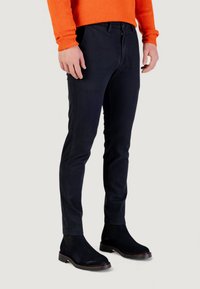 Pantalon noir slim à la texture lisse, avec poches avant et braguette zippée. Associé à des bottines noires montantes et un pull orange.