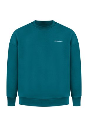 Mand set bagfra iført en turkis sweatshirt med stort hvidt stiliseret grafisk motiv og lille tekst øverst på ryggen, sorte bukser, hænder i lommerne.