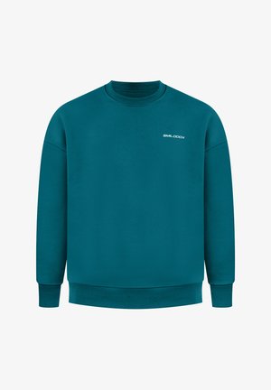 Mann, der mit dem Rücken zur Kamera steht, trägt einen türkisfarbenen Sweatshirt mit großem weißem stilisiertem Grafikmotiv und kleinem Text am oberen Rücken, schwarze Hose, Hände in den Taschen.