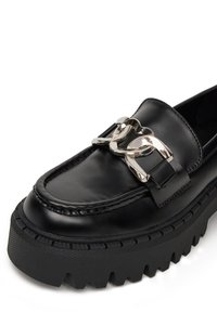 Juodas odinis loaferis su storu guminiu padu. Yra dekoratyvi sidabrinė grandinėlė per viršų ir siūlės detalė palei kraštą.