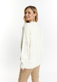 Weißer Strickpullover mit strukturierten Ärmeln, geripptem Saum und floralen Akzenten. Kombiniert mit hellbeigen Hosen, von hinten gezeigt.