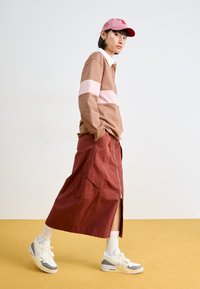 Fustă lungă roșie cu fermoar pe lateral, purtată cu un tricou polo cu mânecă lungă în dungi maronii și roz, și adidași albi cu accente gri.
