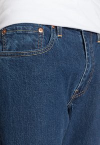 Primer plano de un bolsillo y cinturilla de pantalones vaqueros oscuros de mezclilla azul, con remaches de cobre y costuras naranjas, llevados con una camisa blanca.