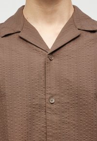 Κοντινό πλάνο καφέ πουκαμίσου με υφή και φαρδιά γιακά, που φοριέται από ένα άτομο, εμφανίζοντας δύο κουμπιά κουμπωμένα στο στήθος.