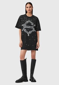 Donna che indossa un abito t-shirt oversize nero con una catena bianca e un design testuale, abbinato a stivali di pelle neri alti fino al ginocchio, in piedi davanti a uno sfondo neutro.