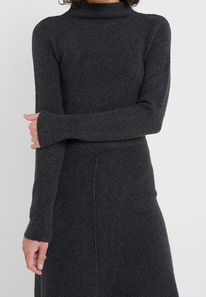 Žena oblečená ve tmavě šedých pletených šatech s dlouhým rukávem a vysokým límcem, s jednou rukou přes druhou, stojící před jednobarevným světlým pozadím.