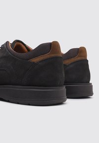 Chaussures en daim noir avec des accents marron, dotées d'un tissu texturé au niveau du col et d'une semelle en caoutchouc avec un léger relief.