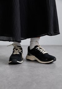 Baskets de sport noires avec dessus en daim et mesh, lacets crème et semelle extérieure texturée. Portées avec des chaussettes blanches montantes et une jupe noire plissée.