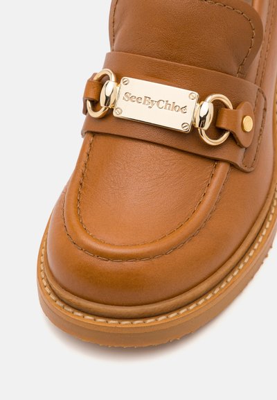 Mocasines de cuero marrón con una placa de logo dorada, doble herraje circular y costura en contraste a lo largo del borde de la suela.