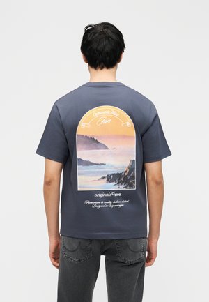 Homme portant un t-shirt gris foncé avec un paysage de coucher de soleil côtier et le texte « Oceanside Bliss Inn » dans le dos, associé à un jean noir.