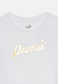 Valge puuvillane T-särk ümmarguste kaelustega, millel on suur kuldne metallist "Puma" logo ja väike Puma kassilogo üleval.