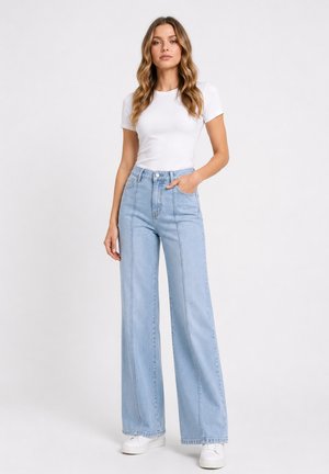 Kobieta ubrana w dopasowany biały t-shirt, jasnoniebieskie jeansy z wysokim stanem i szerokimi nogawkami oraz białe trampki, stojąca z jedną ręką w kieszeni.
