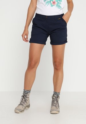 Person trägt marineblaue, hochgekrempelte Shorts, graue Wanderschuhe und graue gemusterte Socken und steht vor einem schlichten hellen Hintergrund.