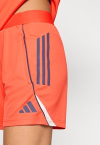 Πορτοκαλί αθλητικό σορτς με λεία υφή, που διαθέτει ναυτικού χρώματος κάθετες ρίγες και λογότυπα. Περιλαμβάνει πλαϊνές τσέπες και αντίθετη λευκή λεπτομέρεια.