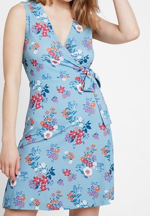 Kvinde iført ærmeløs lyseblå slå-om kjole med flerfarvet blomsterprint og sidebånd, stående mod en hvid baggrund.