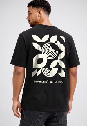 Man in a black t-shirt with bold, abstract white geometric shapes and "NEVERLESS ART STUDIOS" text on the back, standing sideways.