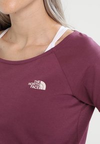 Lyhythihainen, syvän burgundin värinen paita, joka on valmistettu sileästä kankaasta. Paidassa on valkoinen logo vasemmassa rinnassa ja kerrokselliset olkaimet alla.