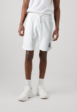 Pantaloni corti sportivi bianchi con elastico in vita e cordino. Presentano tasche laterali e un logo blu scuro. Indossati con sneakers bianche e calzini.