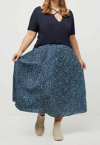 Kvinna iklädd en marinblå kortärmad topp med korslagd halsringning, hållande en blå blommig plisserad midi-skjorta, i kombination med beiga ankelstövlar.