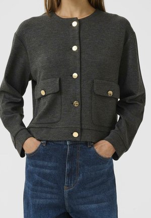 Femme portant une veste boutonnée gris foncé avec deux poches à rabat sur le devant et un jean bleu, les mains dans les poches, sur fond uni.